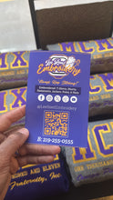 Load image into Gallery viewer, A hand holds a purple business card for Lee Reed Embroidery in front of folded purple and gold embroidered t-shirts. The card includes social media icons, a QR code, and contact information.