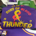 A purple graphic T-shirt with distressed yellow and red text reading 'SONS OF BLOOD & THUNDER' and '1911', photographed on a table with other purple merchandise in a store or event setting.