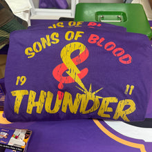 Load image into Gallery viewer, A purple graphic T-shirt with distressed yellow and red text reading 'SONS OF BLOOD &amp; THUNDER' and '1911', photographed on a table with other purple merchandise in a store or event setting.